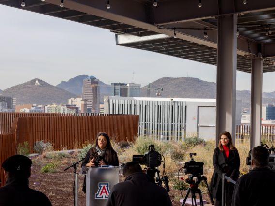 A woman speaks behind a podium with the UArizona logo in front of a backdrop of the Catalina Mountain range
