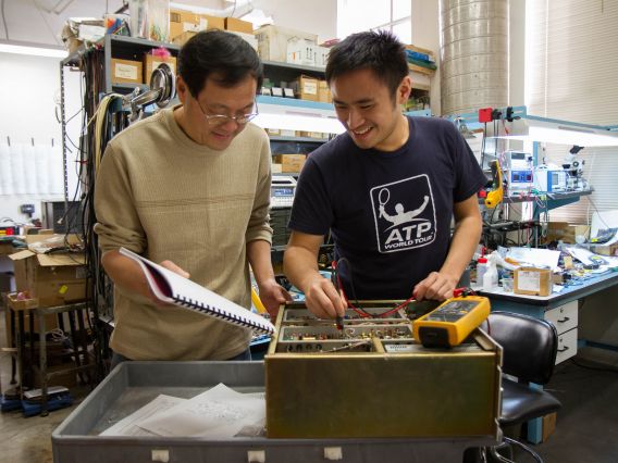 Two men repair lab equipment while consulting a schematic