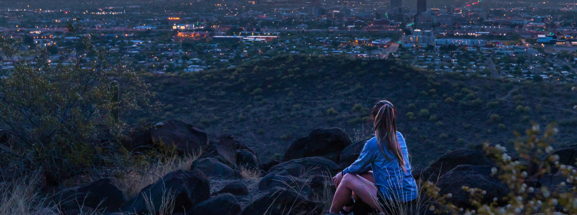 A woman in athlesure clothing sits on Tumamoc Hill looking out over the city of Tucson at dusk