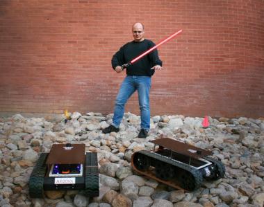 Wolfgang Fink holds a lightsaber and stands in between two of the Mars rovers built in his laboratory.
